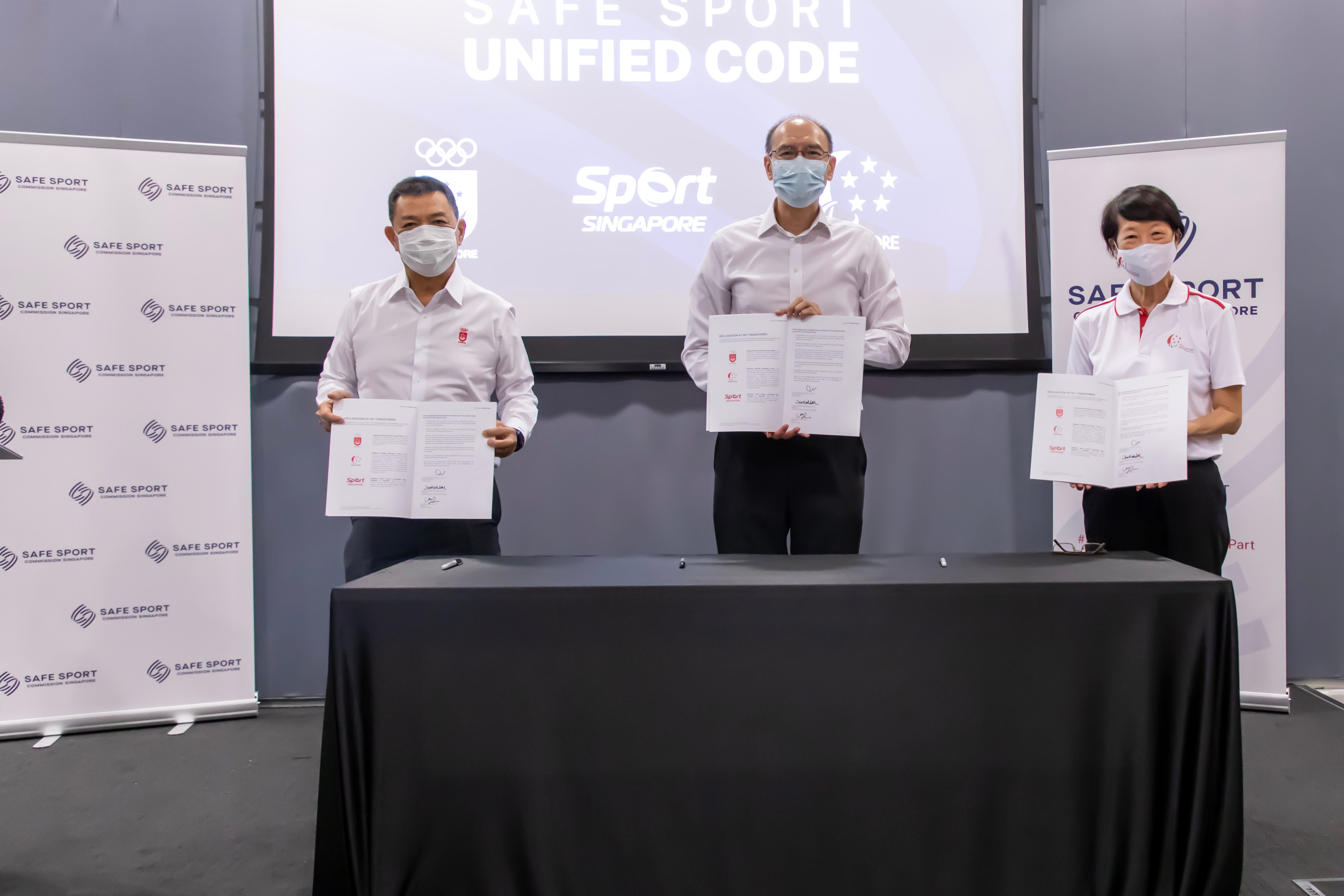 Three masked people wearing masks hold documents behind a black table at a press event, with Safe Sport Singapore branding on screens and banners. The tone is formal and reassuring.